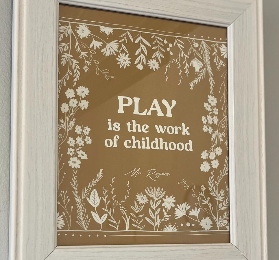 "Play is the Work of Childhood" in white font surrounded by white floral elements, in a white frame.