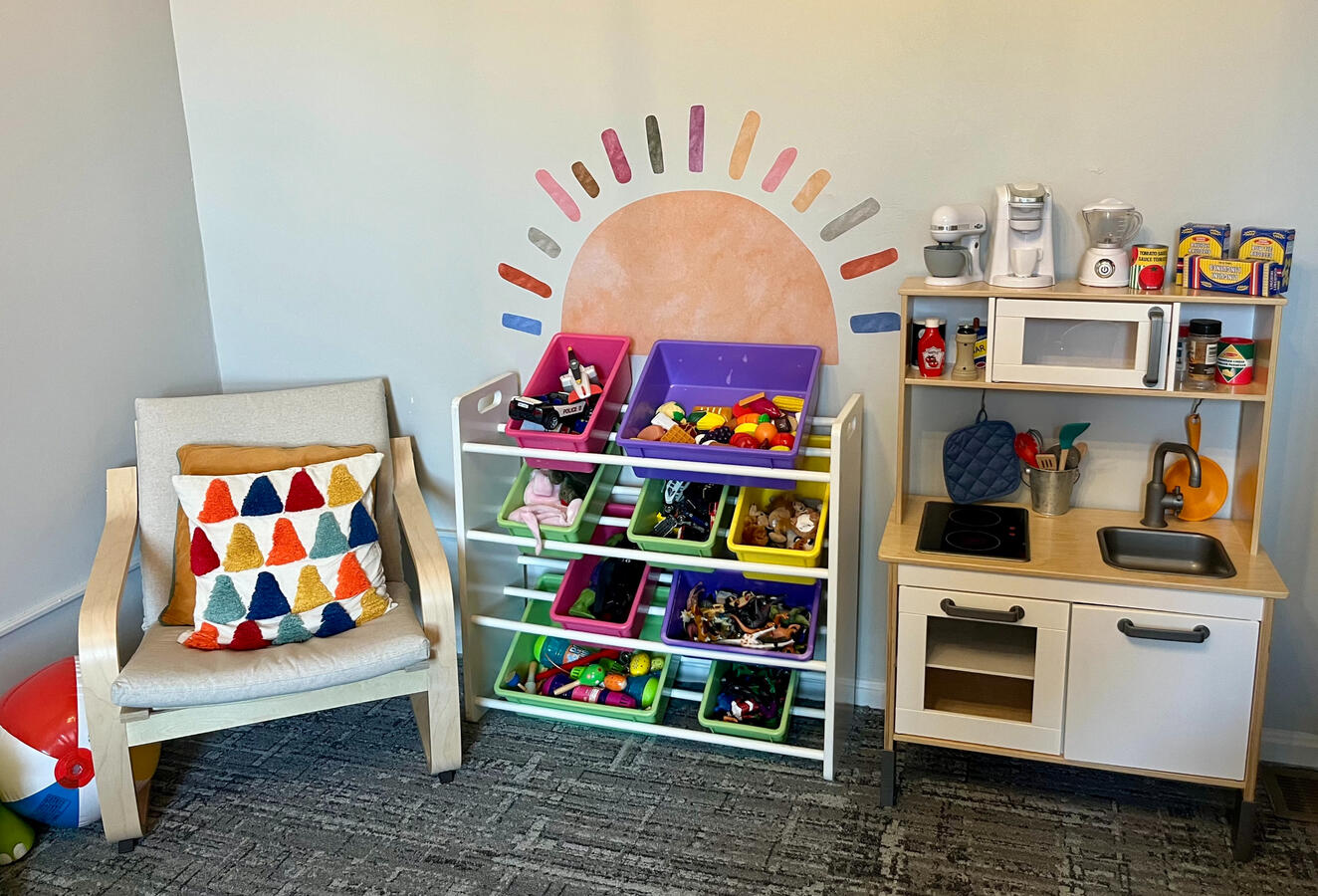 The front wall of the PAYA Playroom, including a seat, a play kitchenette, a shelf of toys, and a sun mural with colorful rays.